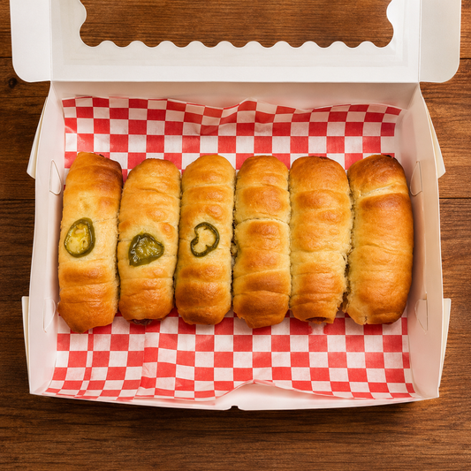 Baked kolaches with jalapeño slices in a box with red and white checkered paper on a wooden surface.