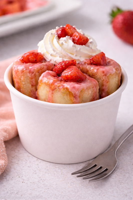 Bowl of strawberry sweet rolls with whipped cream and strawberries on a light surface.