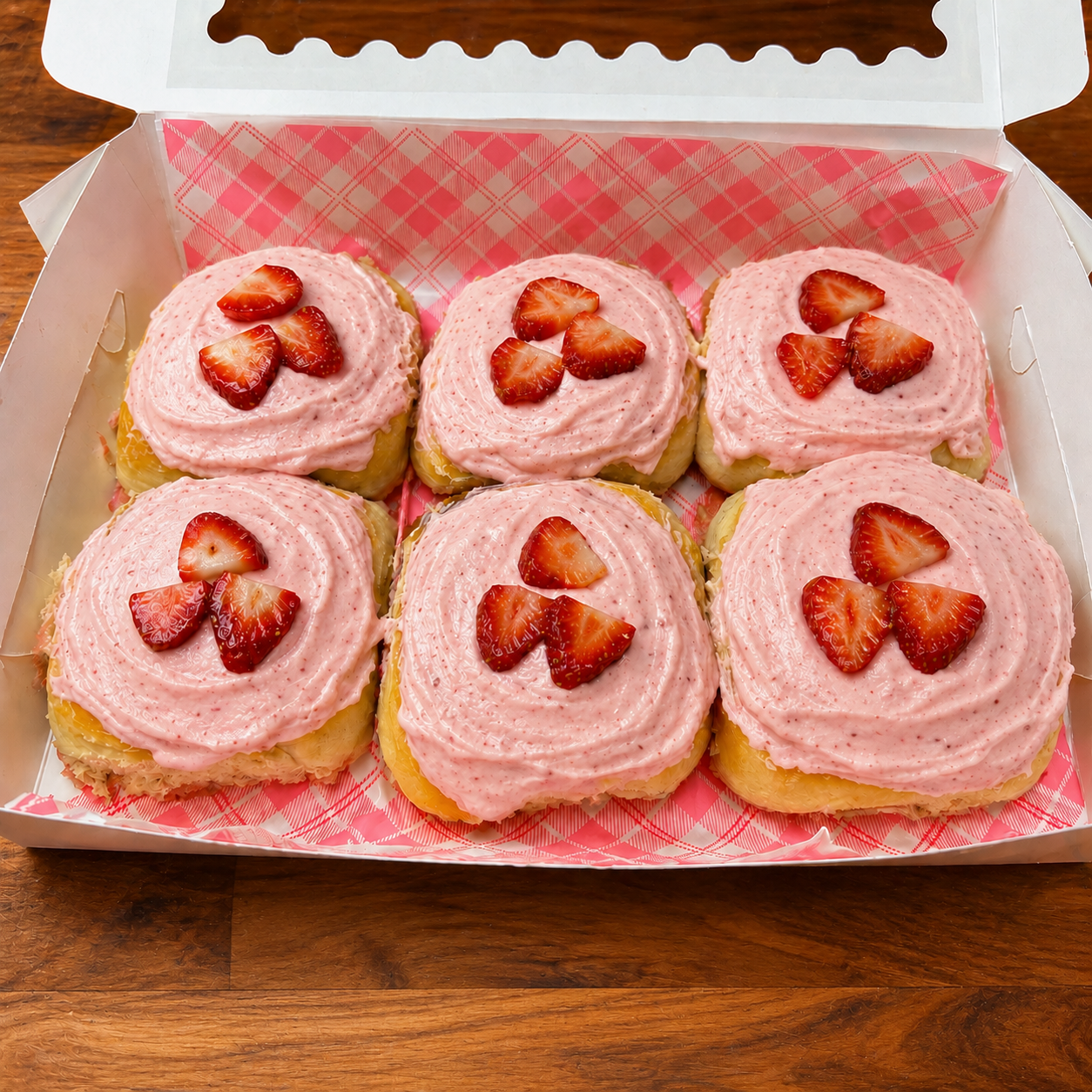Six strawberry scroll pastries with pink frosting and strawberry slices in a box on a wooden surface.