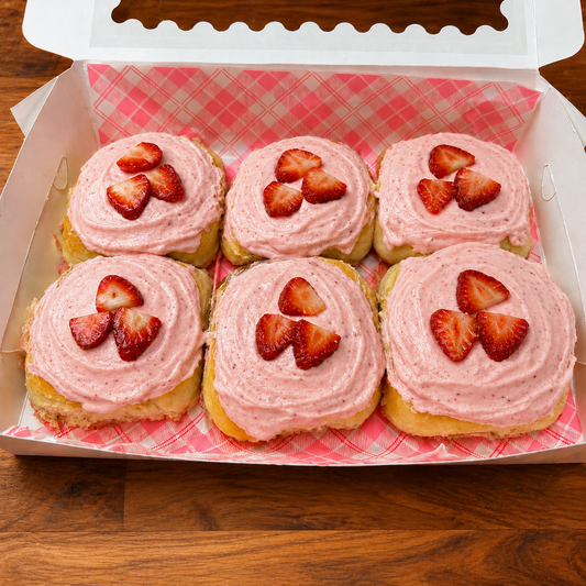 Six strawberry scroll pastries with pink frosting and strawberry slices in a box on a wooden surface.